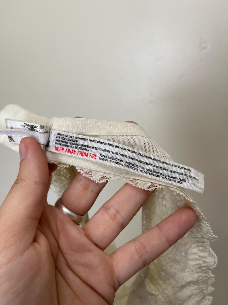 Pu&ograve; includere: Intimo in pizzo bianco sporco con un delicato bordo smerlato. Un'etichetta con istruzioni per la cura e la scritta "KEEP AWAY FROM FIRE" &egrave; visibile. L'indumento &egrave; realizzato in un materiale morbido e leggero.
