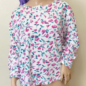 May include: White long-sleeved shirt with a floral print featuring pink and teal flowers on a white background. The shirt has a button closure at the neckline.