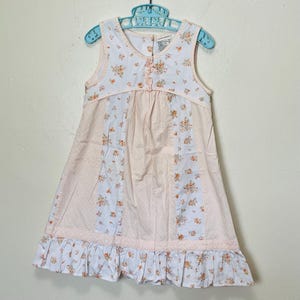 Puede incluir: Vestido rosa claro sin mangas con estampados florales y de lunares. El vestido tiene un dobladillo con volantes y está colgado de una percha de plástico azul. El corpiño tiene un estampado floral y pequeños botones.