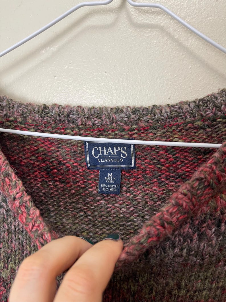 May include: A close-up of a multi-colored sweater with a V-neck. The sweater is a blend of red, green, and brown hues. The label reads "CHAPS CLASSICS" and indicates the sweater is size M, made in China, with 70% acrylic and 30% wool.