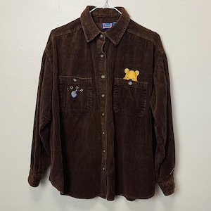 May include: Brown corduroy button-down shirt with long sleeves. Features two front pockets, one with a yellow Winnie the Pooh character peeking out. The other pocket has an embroidered design of a blue honey pot and the word "OOH".