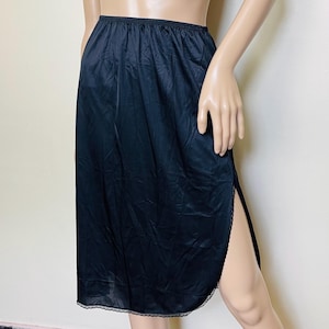 May include: Black knee-length slip skirt with an elastic waistband and a side slit. The skirt has a silky texture and a delicate lace trim along the hemline. This vintage slip skirt is a classic undergarment.