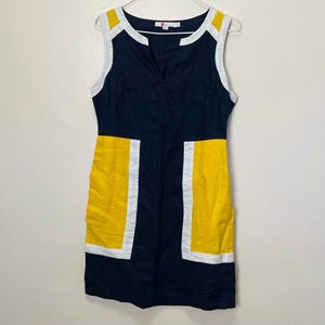 Puede incluir: Vestido sin mangas azul marino con diseño de bloques de color amarillo y blanco. El vestido presenta un escote en V y una silueta recta. Los bloques amarillos están en la parte inferior delantera, contorneados en blanco.