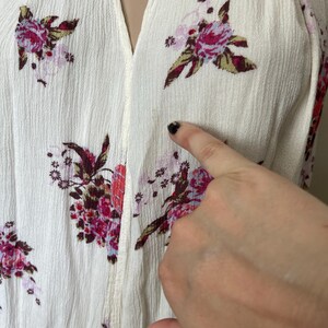 Op de afbeelding: Cr&egrave;mekleurige top met een V-hals en een bloemenpatroon in roze, rode en groene tinten. De stof heeft een getextureerde uitstraling. De afbeelding toont een close-up van de top.