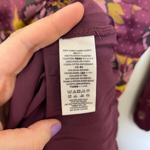 May include: A burgundy floral print dress with a gathered neckline and long sleeves. The fabric is 100% polyester and the lining is 100% rayon. The care instructions are: machine wash cold with like colors, use only non-chlorine bleach, tumble dry low, low iron as needed, may be dry cleaned.