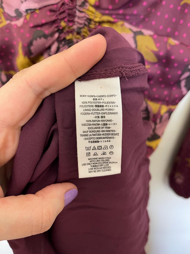May include: A burgundy floral print dress with a gathered neckline and long sleeves. The fabric is 100% polyester and the lining is 100% rayon. The care instructions are: machine wash cold with like colors, use only non-chlorine bleach, tumble dry low, low iron as needed, may be dry cleaned.