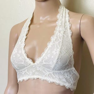 Pu&ograve; includere: Bralette avorio con stile a fascia. Il bralette &egrave; realizzato in delicato pizzo con bordo smerlato. Il design presenta uno scollo a V e un cinturino all'americana che si allaccia dietro il collo. Il motivo in pizzo include dettagli floreali e geometrici.