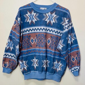 May include: A blue, white, and brown patterned sweater with a crew neck and long sleeves. The sweater features a geometric design with star and zigzag patterns. The cuffs and neckline are a solid light blue colour. The tag reads "SALT AIR".