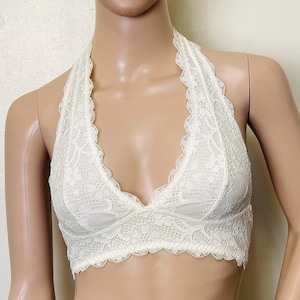 Puede incluir: Bralette color marfil con diseño halter y delicado encaje floral. El bralette presenta un borde festoneado a lo largo del escote y la parte inferior. El diseño es de estilo suave y femenino.