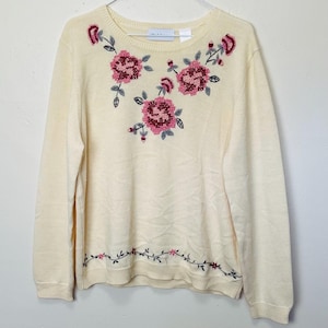 May include: A cream-colored sweater with embroidered pink and gray flowers. The sweater has a crew neck and long sleeves.