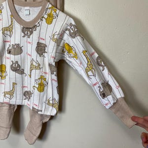 May include: A white long-sleeved pajama top and matching pants set. The top features a pattern of various cartoon animals, including giraffes and lions, with vertical stripes. The cuffs and neckline are tan.
