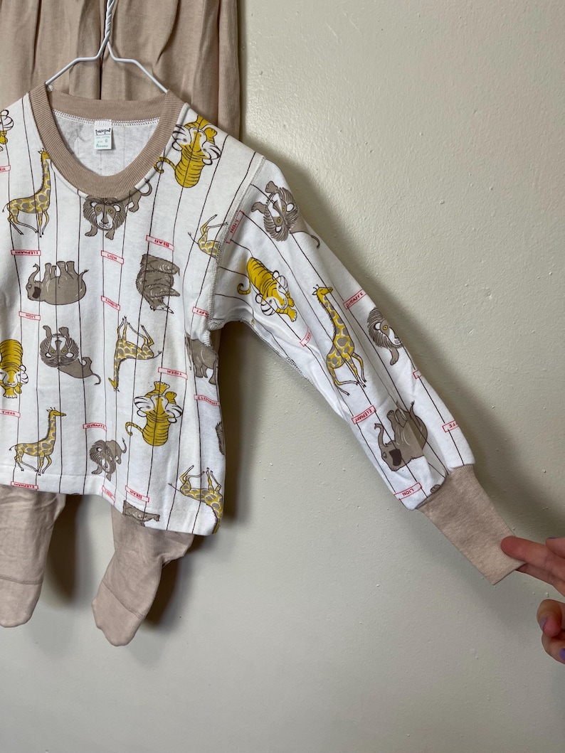 May include: A white long-sleeved pajama top and matching pants set. The top features a pattern of various cartoon animals, including giraffes and lions, with vertical stripes. The cuffs and neckline are tan.