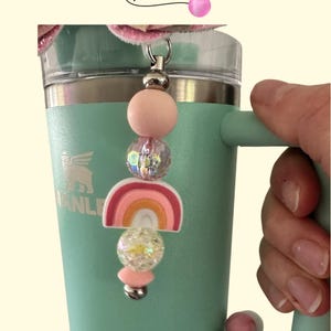 Puede incluir: Un dije de cuentas con un diseño de arcoíris y cuentas rosas y transparentes está unido a un vaso de color turquesa. El dije tiene un cierre de metal plateado. El vaso tiene un asa y la palabra "Stanley" es visible.