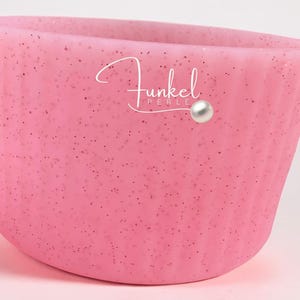 Op de afbeelding: Een roze, met glitter bedekte container met een gebogen vorm. De container heeft het woord "Funkel" in cursief schrift, met een parel accent. De container is lichtroze van kleur met kleine, donkere glitterdeeltjes.