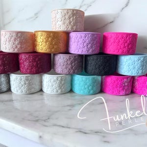 Può includere: Una collezione di fasce in silicone cilindriche colorate, ciascuna ornata da un motivo floreale. Le fasce sono impilate su una superficie di marmo, mostrando una varietà di colori tra cui rosa, oro, viola e blu. Il marchio "Funkel Perle" è visibile.