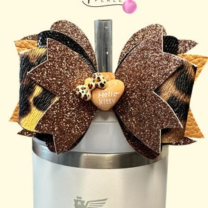 Könnte beinhalten: Eine dekorative Schleife mit glitzerndem braunen Design, Leopardenmuster und einem herzförmigen Hello Kitty-Ornament. Die Schleife ist an einem weißen Stanley-Becher befestigt, ideal für Getränke.