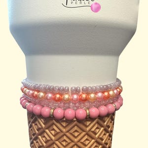 Op de afbeelding: Een stapel roze kralenarmbanden op een goudkleurige, getextureerde basis. De armbanden variëren in tint, van lichtroze tot een donkerdere roos, met enkele heldere kralen en gouden accenten. De bovenkant is wit, met de tekst "Funkel Perle".