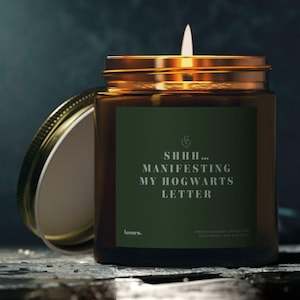 Puede incluir: Una vela encendida en un tarro de cristal con tapa dorada y etiqueta verde. La etiqueta dice "SHHH... MANIFESTING MY HOGWARTS LETTER". La vela está sobre una superficie oscura y texturizada, con la tapa ligeramente entreabierta.