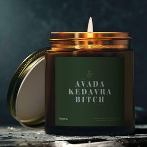 Könnte beinhalten: Eine brennende Kerze in einem Glas mit goldenem Deckel und grünem Etikett. Auf dem Etikett steht "AVADA KEDAVRA BITCH" in weißer Schrift. Die Kerze steht auf einer dunklen Oberfläche mit dunklem Hintergrund.