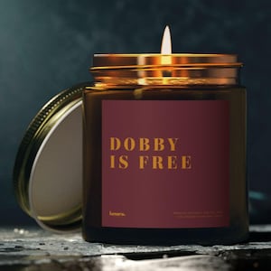 May include: A lit candle in a glass jar with a gold lid. The jar has a burgundy label with the text "DOBBY IS FREE" in gold lettering. The candle is on a dark, textured surface.