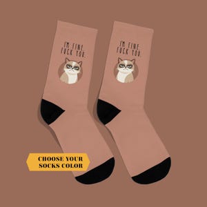May include: A pair of socks with a light brown background, black heels and toes. Each sock features a grumpy cat graphic with the text "I'M FINE. FUCK YOU." above the cat. A yellow banner at the bottom says "CHOOSE YOUR SOCKS COLOR."
