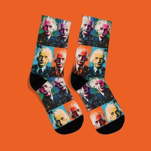 May include: A pair of black socks with a colourful pop art design featuring multiple portraits of Albert Einstein. The portraits are set against various coloured backgrounds, including blue, orange, and yellow. The socks have black heels and toes.