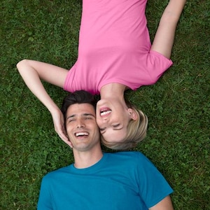 May include: A top-down shot of two people lying on green grass, smiling. One person wears a pink t-shirt and jeans, the other a blue t-shirt. The woman's head is near the man's, both laughing.