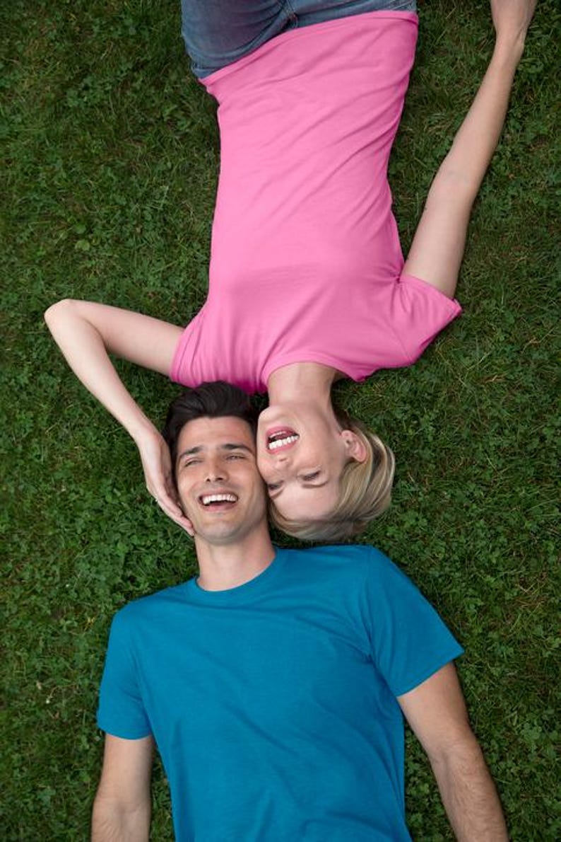 May include: A top-down shot of two people lying on green grass, smiling. One person wears a pink t-shirt and jeans, the other a blue t-shirt. The woman's head is near the man's, both laughing.