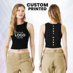 May include: Black crop top with the text "YOUR LOGO HERE" on the front and "TEXT HERE" on the back. The words "CUSTOM PRINTED" are at the top. The model is wearing khaki pants.
