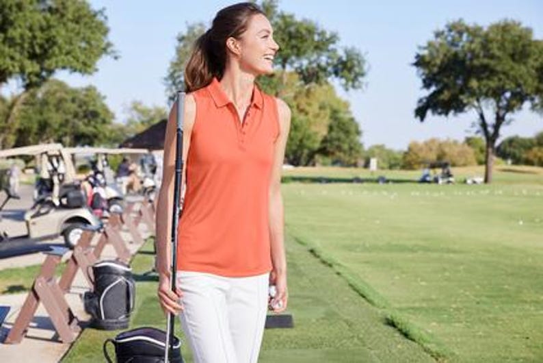 Puede incluir: Una mujer con un polo naranja sin mangas y pantalones blancos est&aacute; en un campo de golf, sosteniendo un palo de golf. Se ven carritos de golf y &aacute;rboles en el fondo en un d&iacute;a soleado.