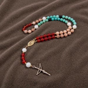 May include: A multi-coloured rosary featuring a silver crucifix and a gold-coloured connector. The beads are a mix of red, white, pink, and turquoise. The rosary is displayed on a brown, textured surface.