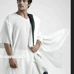 Könnte beinhalten: Eine weiße Leinen-Tunika mit einem langen, fließenden Design. Die Tunika hat einen V-Ausschnitt und kurze Ärmel. Das Model trägt eine weiße Hose und einen schwarzen Schal.