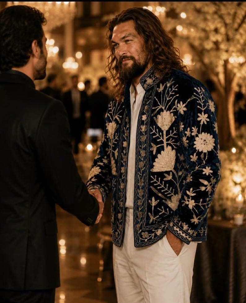Pu&ograve; includere: Una giacca in velluto blu profondo con intricati ricami floreali dorati, indossata sopra una camicia bianca abbottonata e pantaloni bianchi. La giacca presenta un colletto alto e maniche lunghe, con cuciture dorate sul davanti e sulle maniche.