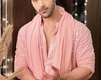 Può includere: Un kurta rosa chiaro con ricami intricati e una sciarpa abbinata. L'outfit è accessoriato con un orologio d'oro, una catena d'oro e un braccialetto di perline. La persona ha i capelli scuri e una barba ben curata.