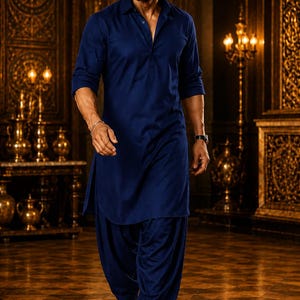 May include: A man in a navy blue kurta pajama, a traditional South Asian outfit, walks forward. The outfit includes a long tunic and loose-fitting pants. He wears black dress shoes. The background features ornate gold decor and lighting.