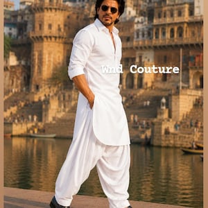 Könnte beinhalten: Ein Mann in einem weißen Kurta- und Salwar-Anzug mit schwarzen Schuhen steht auf einer Steinoberfläche. Der Text "Wind Couture" ist sichtbar. Der Hintergrund zeigt Wasser und Gebäude.