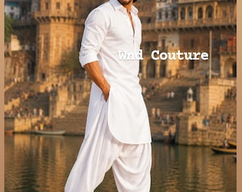 Può includere: Un uomo indossa un completo kurta e salwar bianco, con scarpe nere, in piedi su una superficie di pietra. Il testo "Wind Couture" è visibile. Lo sfondo presenta acqua ed edifici.