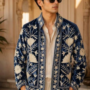 Pu&ograve; includere: Una giacca in velluto blu profondo caratterizzata da intricati ricami floreali color crema sui pannelli frontali, sul colletto e sui polsini. La giacca &egrave; indossata sopra una camicia beige chiaro abbottonata e pantaloni beige chiaro, con occhiali da sole.