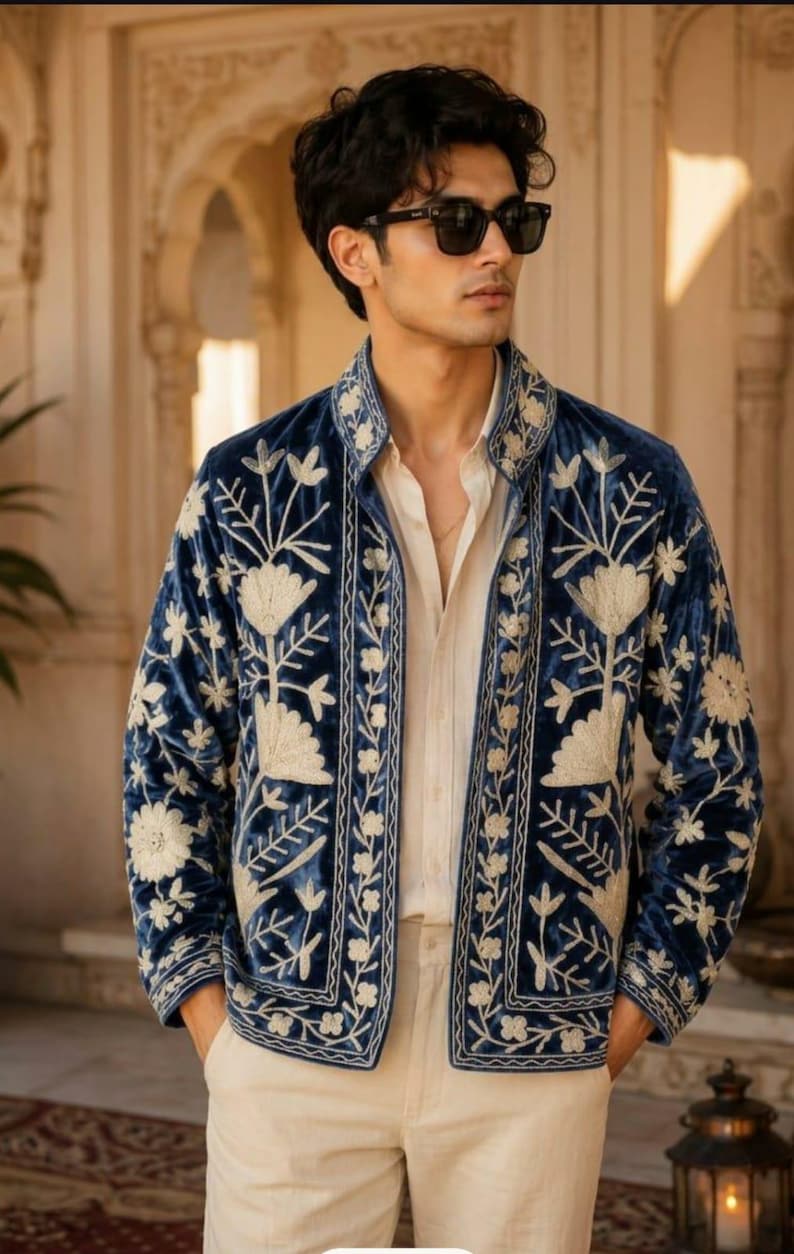 Pu&ograve; includere: Una giacca in velluto blu profondo caratterizzata da intricati ricami floreali color crema sui pannelli frontali, sul colletto e sui polsini. La giacca &egrave; indossata sopra una camicia beige chiaro abbottonata e pantaloni beige chiaro, con occhiali da sole.