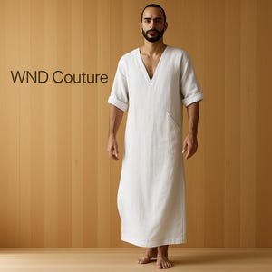 Op de afbeelding: Een lange, witte jurk met een V-hals en korte mouwen. De jurk heeft een enkele zak en is gemaakt van een lichte stof. De tekst "WND Couture" is links zichtbaar. De jurk komt tot de enkels.