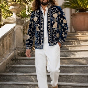 Pu&ograve; includere: Una giacca in velluto blu navy con intricati ricami floreali dorati sui pannelli frontali e sul colletto, indossata sopra una camicia bianca abbottonata e pantaloni bianchi, con mocassini in camoscio beige e occhiali da sole scuri.