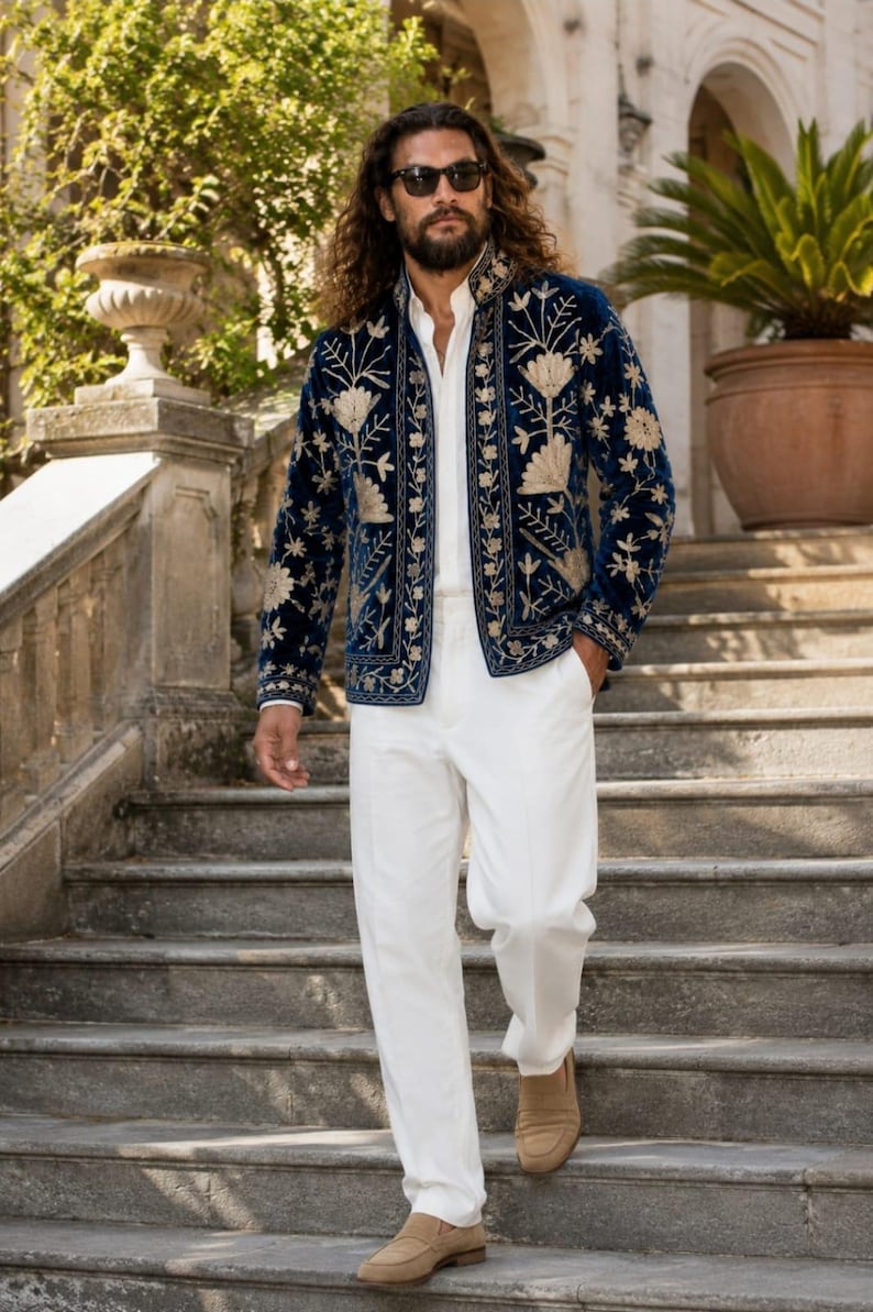 Pu&ograve; includere: Una giacca in velluto blu navy con intricati ricami floreali dorati sui pannelli frontali e sul colletto, indossata sopra una camicia bianca abbottonata e pantaloni bianchi, con mocassini in camoscio beige e occhiali da sole scuri.