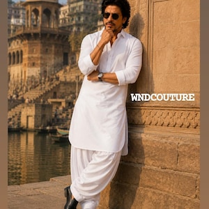 May include: A man in a white kurta pajama, leaning against a stone wall. He wears black shoes and a watch. The image includes the text "WNDCOUTURE". The background features a cityscape with a river and buildings.