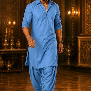 May include: A man in a light blue kurta pajama, a traditional South Asian outfit, walks forward. The outfit includes a collared shirt and loose-fitting pants. He wears black dress shoes and a watch. The background features ornate decor and warm lighting.