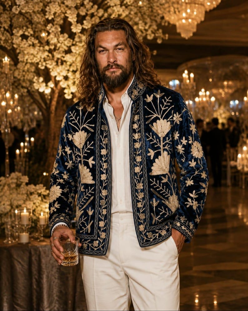 Pu&ograve; includere: Una giacca in velluto blu profondo con intricati ricami floreali dorati sui pannelli frontali e sui polsini, indossata sopra una camicia bianca abbottonata e abbinata a pantaloni bianchi, creando un completo formale con capelli lunghi.