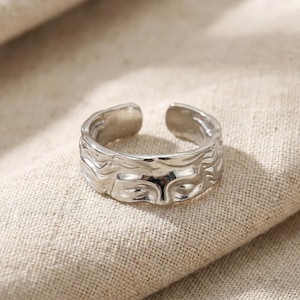 Könnte beinhalten: Ein silberfarbener Metallring mit offenem Banddesign und einer texturierten, wellenförmigen Oberfläche, die an fließendes Wasser oder organische Wellen erinnert. Der Ring hat eine polierte, reflektierende Oberfläche und eine breite, unregelmäßige Form.