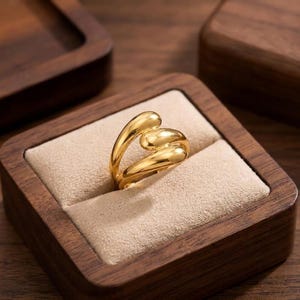 Könnte beinhalten: Ein goldener Ring mit glatter, polierter Oberfläche und einem fließenden, offenen Design, das an drei miteinander verbundene, abgerundete Tropfenformen erinnert. Der Ring befindet sich in einer quadratischen Holzschatulle mit beigem Innenfutter.