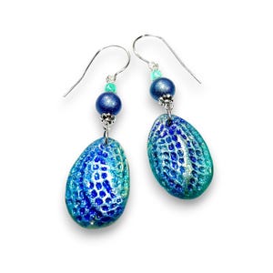 Puede incluir: Un par de pendientes de color azul y verde en forma de lágrima con una superficie texturizada. Los pendientes tienen un gancho plateado y una pequeña perla azul.