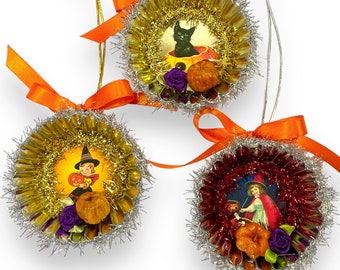 May include: Three round Halloween ornaments featuring vintage-style illustrations of a black cat, a child in a witch hat, and a child in a red cape. Each ornament is framed by metallic tinsel, orange ribbon bows, small pumpkins, and purple flowers.