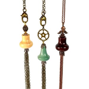 May include: Three necklaces with wooden pendant charms. The charms are shaped like bells and have different colors and finishes. The necklaces have gold chains and tassels.